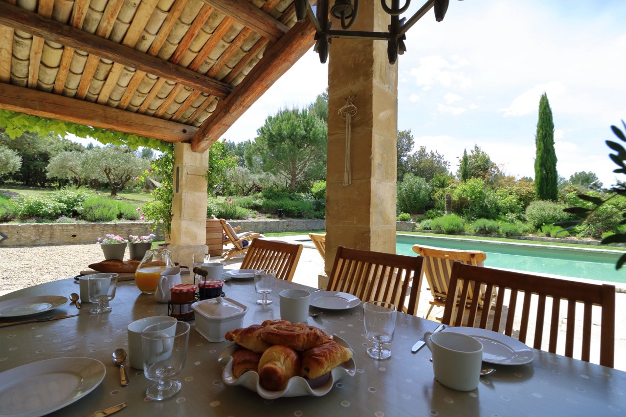 Outdoor breakfast table set with croissants and coffee under a pergola, overlooking a serene garden and swimming pool, in bright, sunny weather.